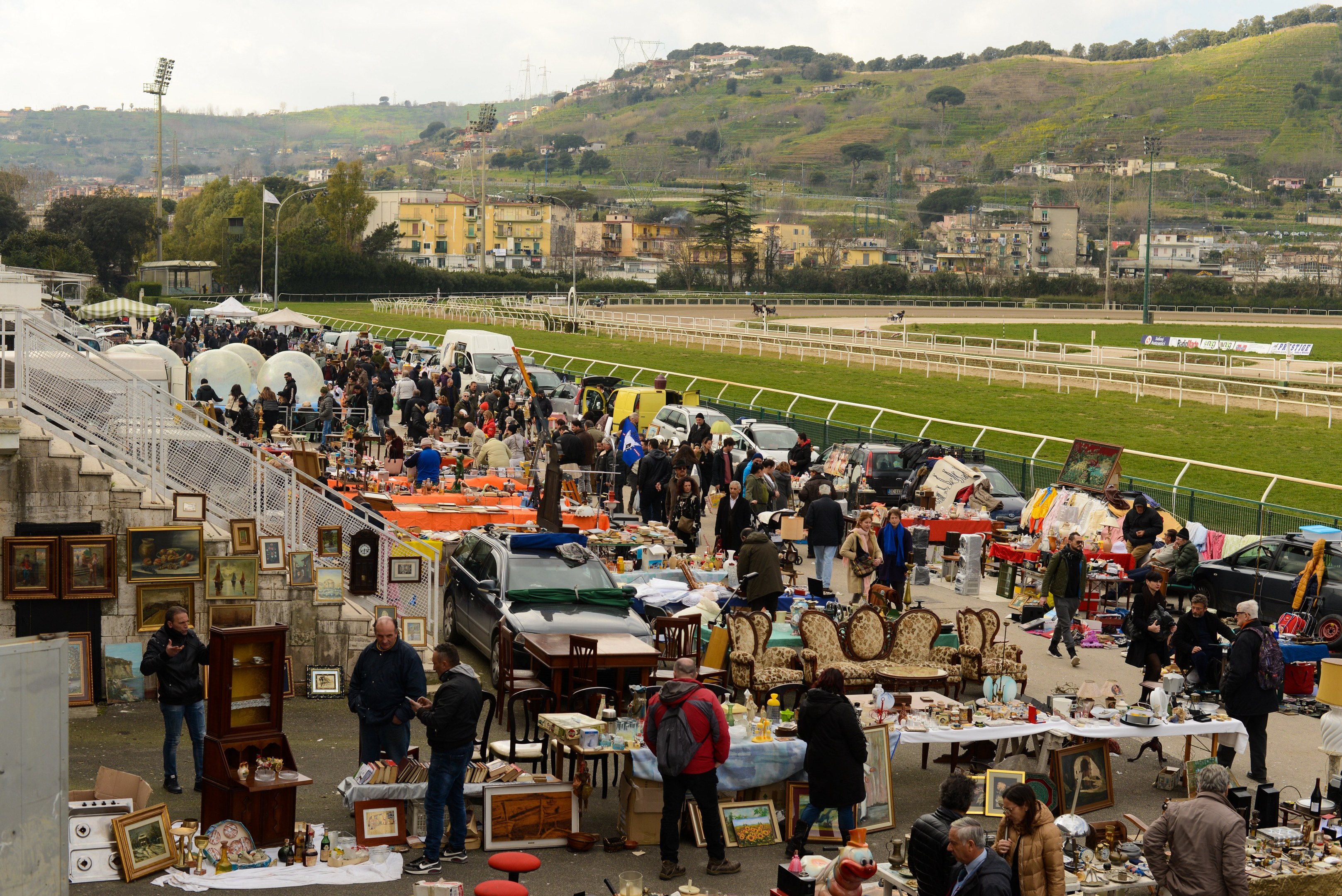 Eine große Gruppe von Menschen auf einem Flohmarkt mit Tischen, auf denen Gegenstände wie Foto Rahmen und Stühle ausgelegt sind, umgeben von parkenden Fahrzeugen, Geländern, Treppen, Bäumen, Gebäuden, Laternenmasten, Hügeln und einem bewölkten Himmel.