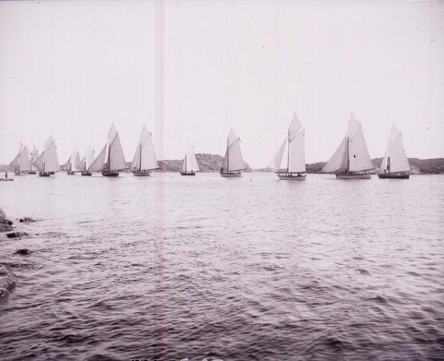 Schwarzes und weißes Foto von Segelbooten auf dem Wasser mit Hügeln im Hintergrund und einem klaren Himmel, beschriftet als New York Yacht Club.