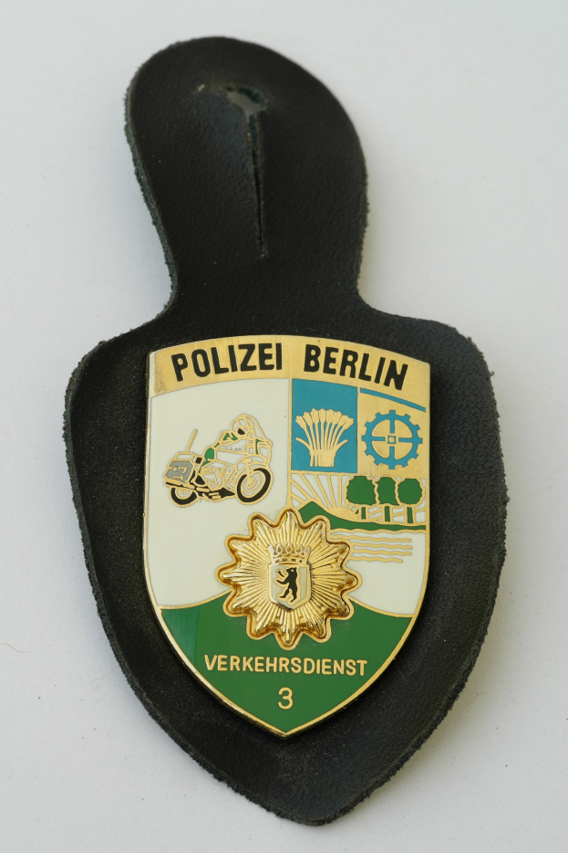 Ein schildförmiges Polizeibadge mit "Polizei Berlin" in fetter schwarzer Schrift auf einem blauen Hintergrund, das ein weißes Sternchen in der Mitte und einen weißen Rand hat.