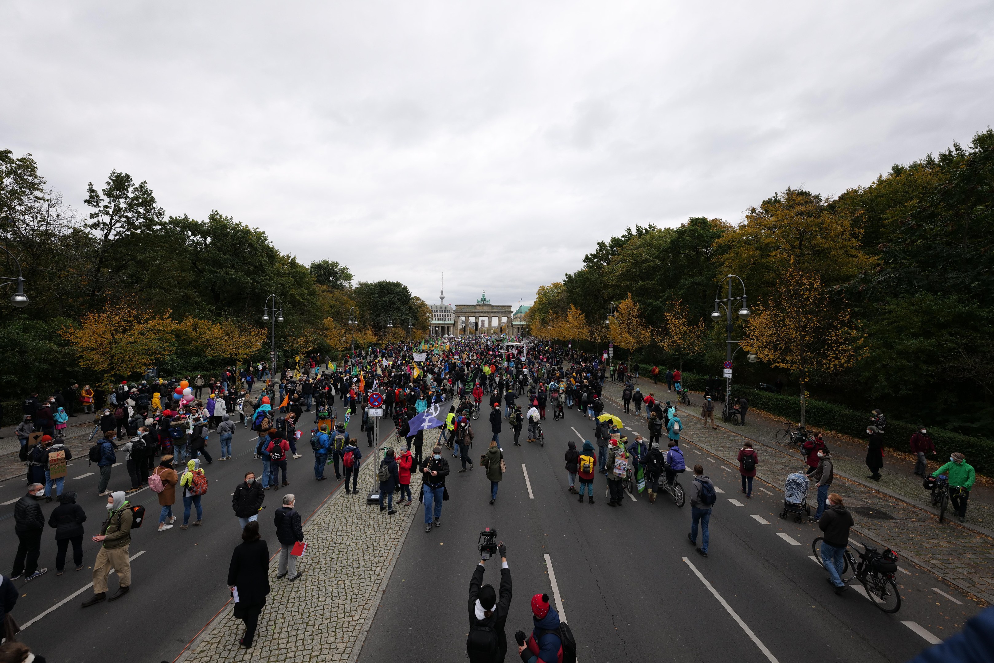 Eine große Gruppe von Menschen marschiert auf einer von Bäumen gesäumten Straße mit Laternenmasten, mit Kameras in der Hand, vor einem Gebäude unter einem klaren Himmel.