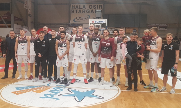 Gruppe von Männern und Kindern auf einem Basketballfeld mit Schildern "Hala Osir Starga", sitzende Zuschauer, ein Basketballkorb, Metallstangen, Deckenbeleuchtung und eine Wand im Hintergrund.