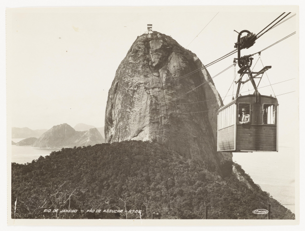 Schwarze und weiße Fotografie einer Seilbahn, die einen Berg hinauffährt, mit einem sichtbaren Passagier im Inneren, umgeben von Bäumen, Hügeln und Wasser, und Text am unteren Rand.