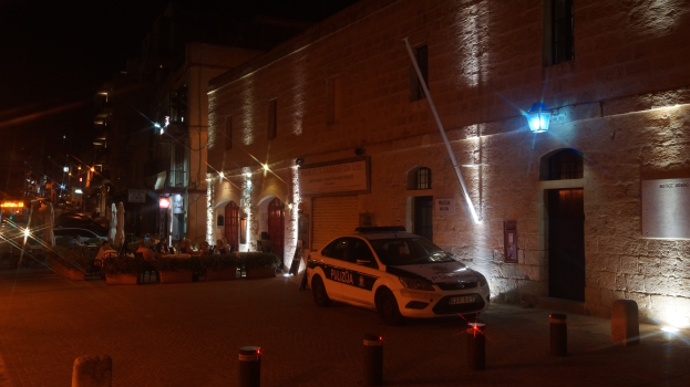 Ein Polizeiauto auf einer Straße bei Nacht, umgeben von Pollern, Topfpflanzen, Menschen auf Stühlen, anderen Fahrzeugen, Gebäuden mit Fenstern, Schildern und Straßenlaternen unter einem bewölkten Himmel.