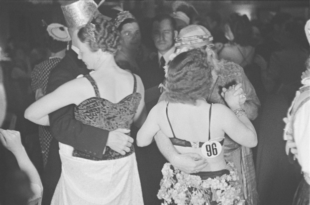 Schwarzes und weißes Foto einer Gruppe von Menschen, die auf einer Party tanzen, mit einer Frau in einem Kleid und Hut in der Mitte.