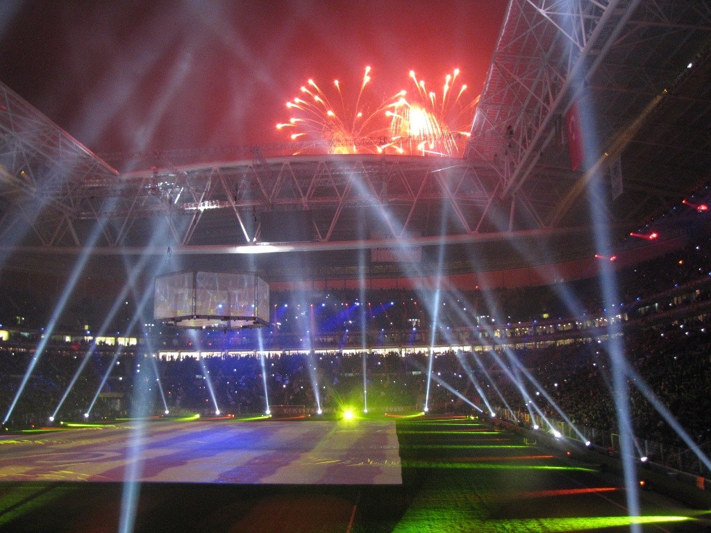 Olympiastadion in London bei Nacht, beleuchtet von hellen Lichtern und Feuerwerken, mit einem großen Bildschirm im Vordergrund und einer Menge drinnen.