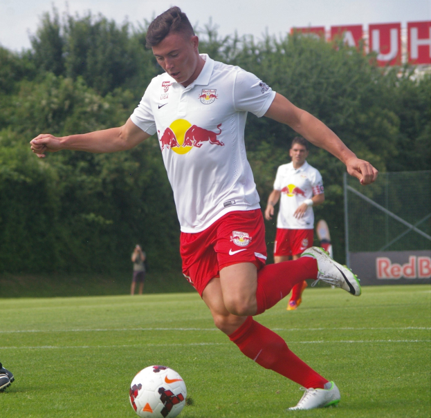 Professioneller Fußballspieler, der einen Ball auf einem grünen Feld mit Bäumen und einem klaren blauen Himmel im Hintergrund kickt, RB Leipzig-Logo sichtbar.