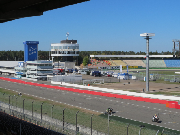 Gruppe von Motorradfahrern auf einer Rennstrecke, umgeben von einer Absperrung, Fahrzeugen, einem Gebäude, einem Schild, einer Tribüne, Bäumen und einem bewölkten Himmel.