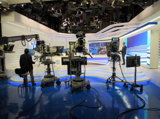 Ein TV-Studio mit mehreren bodengestützten Kameras, einer sitzenden Person im Vordergrund und einigen stehenden Individuen im Hintergrund, das Deckenleuchten aufweist, die auf eine Livesendung hindeuten.