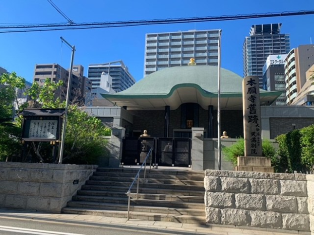 Großes grünoberes Gebäude in städtischer Umgebung mit umliegenden Treppen, Geländern, Beschilderung, Grünflächen und einer Straße darunter, vor einem klaren blauen Himmel.