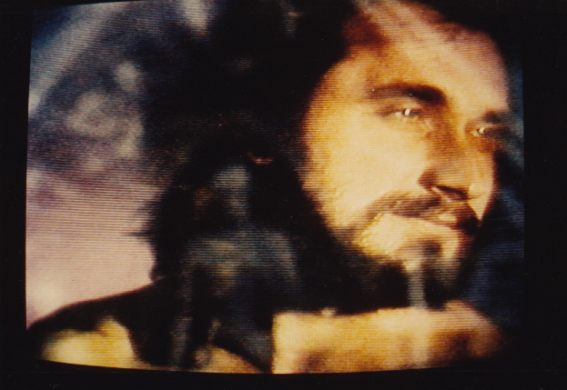 Ein Mann mit dunklem Bart und langen dunklen Haaren, der ein weißes Hemd trägt, erscheint auf einem Fernsehbildschirm mit einem ernsten Gesichtsausdruck und direktem Blick.