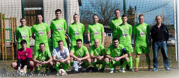 Eine Gruppe von Männern in grünen T-Shirts steht auf einem Fuballfeld, einige hocken, mit einem Ball und einem Torpfosten; der Hintergrund umfasst Bäume, Gebäude und Fahrzeuge.