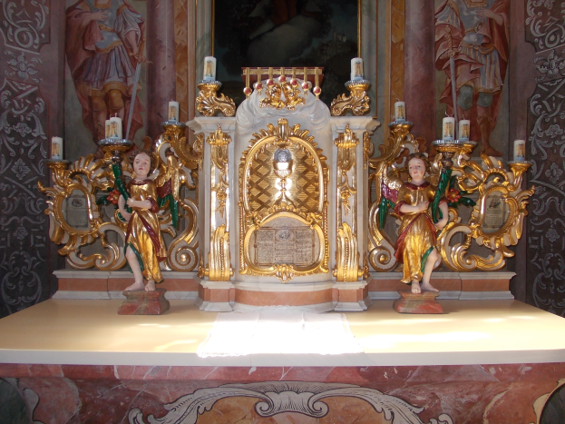 Ein Altar in einer Kirche mit Skulpturen von Menschen und Kerzen auf Ständern, mit einem Gemälde an der Wand dahinter.