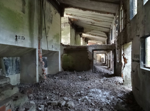 Altes Gebäude mit erheblichen strukturellen Schäden, zerbrochene Fenster, verstreute Trümmer, freiliegende Rohre und Schutt auf dem Boden und den Wänden.