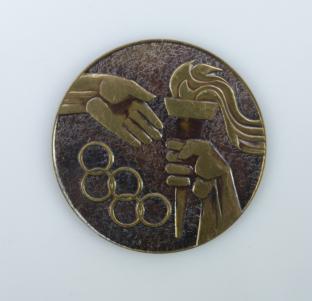 Eine runde Bronzemedaille mit detaillierten Verzierungen und den Olympischen Ringen vor einem weißen Hintergrund.