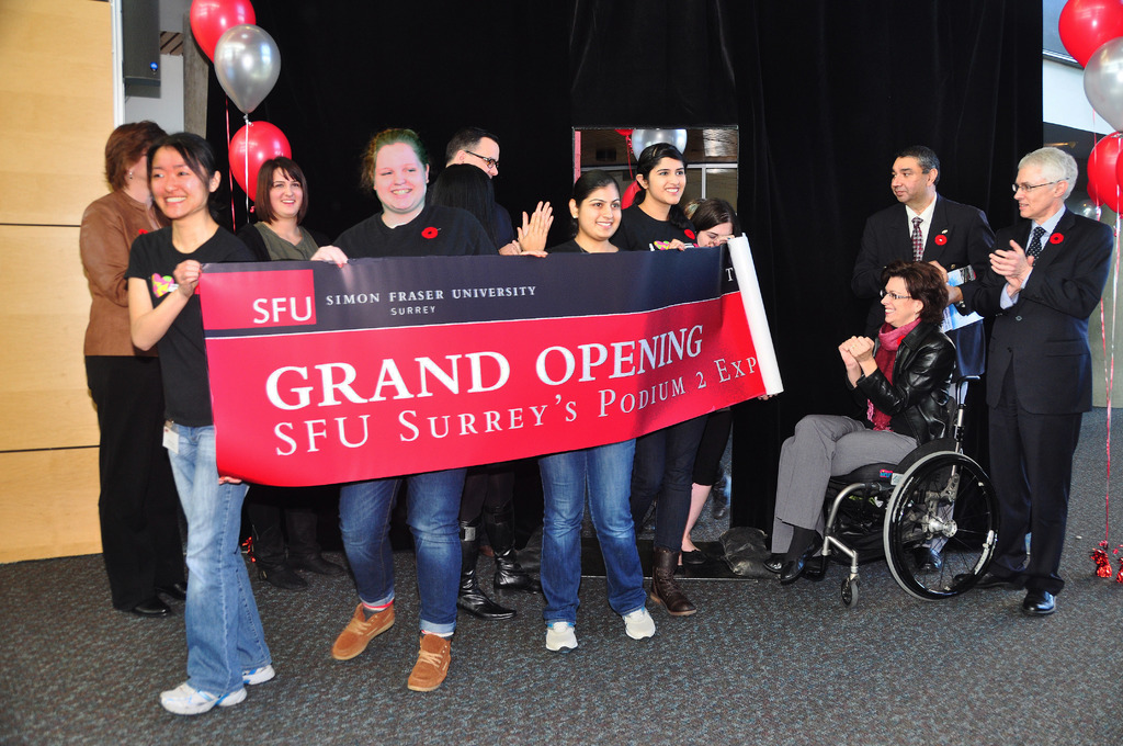 Eine Gruppe von Frauen hält ein Banner, das ein Ereignis ankündigt, mit zwei klatschenden Männern daneben und einer Frau im Rollstuhl, die aufgeregt aussieht, vor einem schwarzen Vorhang und Ballons.