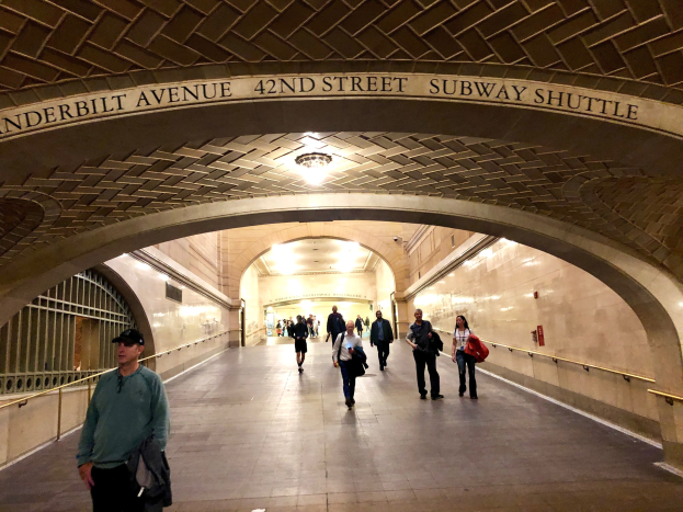 Eine Gruppe von Menschen geht durch eine belebte U-Bahn-Station in New York City mit Deckenleuchten, einem Geländer auf der linken Seite und Text an den Wänden.