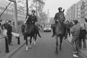 Zwei Polizeibeamte reiten auf Pferden eine Straße entlang, während ein Auto neben ihnen fährt, und Menschen stehen auf den Gehwegen und Gebäude, Bäume und ein klarer Himmel sind im Hintergrund zu sehen.
