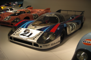 Ein Porsche 917K Le Mans-Rennwagen wird in einem Museum ausgestellt, umgeben von anderen Sportwagen und Menschen, mit einer Wand im Hintergrund und einem Blatt Papier mit Text auf der linken Seite.
