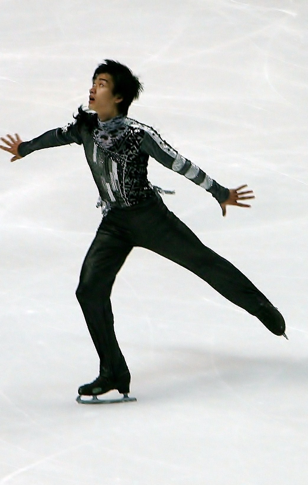Männlicher Eiskunstläufer in schwarzer Kleidung gleitet über eine Eisfläche und führt eine Choreographie aus.