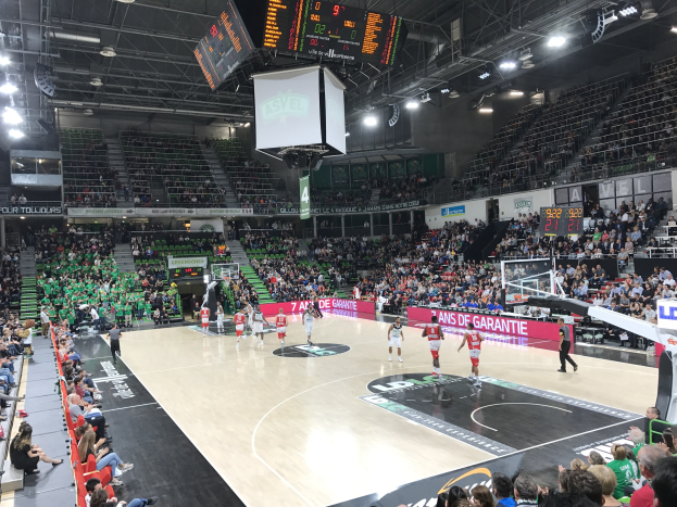 Basketballspiel in einer großen Arena mit Publikum, Anzeigetafeln mit "Asvel vs Galatasaray" und Decke mit Lampen und Bildschirm.