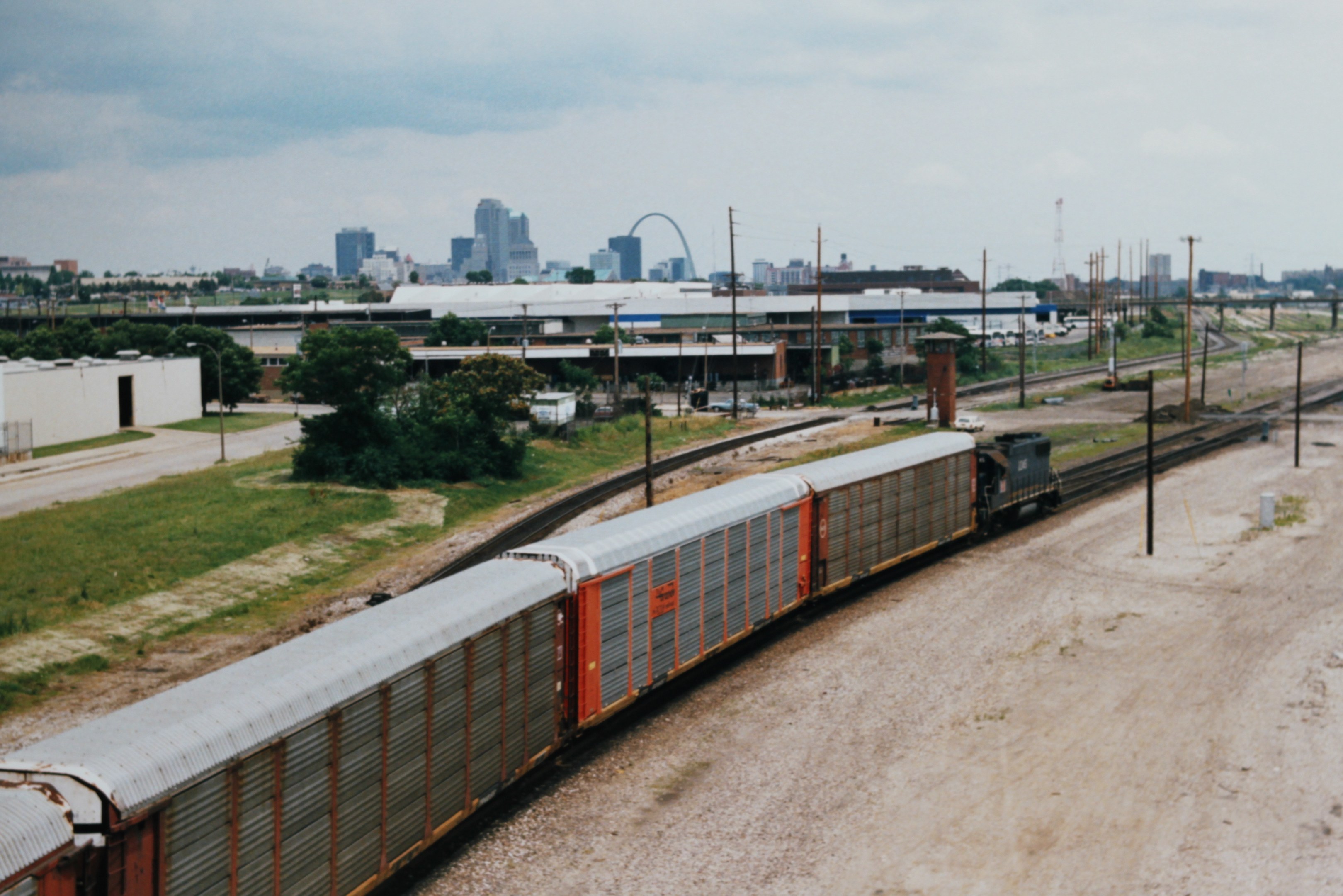 Güterzug fährt auf Gleisen neben einer Stadtlandschaft mit Strommasten, Kabeln, Gebäuden, Bäumen, Gras und bewölktem Himmel.