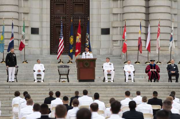 Gruppe von Menschen in weißen Uniformen auf den Stufen eines Gebäudes mit einem Podium, das von Fahnen flankiert ist, vor einem großen Gebäude mit einer Tür während einer Ehrenzeremonie.