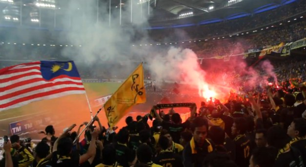 Eine große Menge Menschen in einem Stadion mit Fackeln in der Luft, einige halten Fahnen und eine Person hält eine Kamera, mit Rauch im Hintergrund und Schildern mit der Aufschrift "Malaysia vs Malaysia Live Streaming".