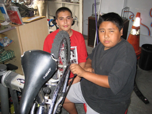 Zwei Personen reparieren ein Fahrrad an einem unordentlichen Schreibtisch in einem Raum mit Fahrrädern, Ersatzteilen und Lagerbeständen im Hintergrund.