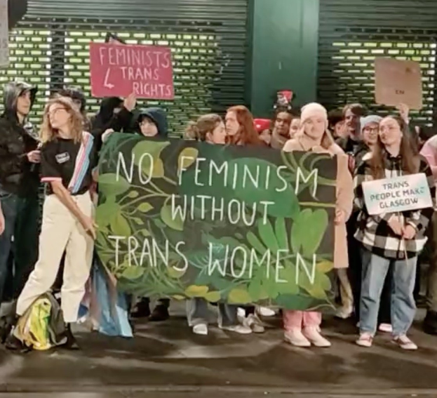Gruppe von Menschen mit einem Banner, auf dem "Kein Feminismus ohne Transfrauen" steht, und Schildern an einemöffentlichen Platz mit einer Wand und einem Rollladen im Hintergrund.