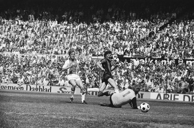 Schwarze-weiß-Fotografie von Männern, die auf einem Feld Fussball spielen, mit Zuschauern im Hintergrund und einer "Bundesliga 1978-79 Ajax 1-1 Hannover 96"-Tafel.