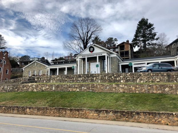 Steinmauer mit parkenden Autos, benachbarten Gebäuden mit Fenstern und Türen, Bäumen und einem bewölkten Himmel, beschriftet als Weltkulturerbe-Museum.