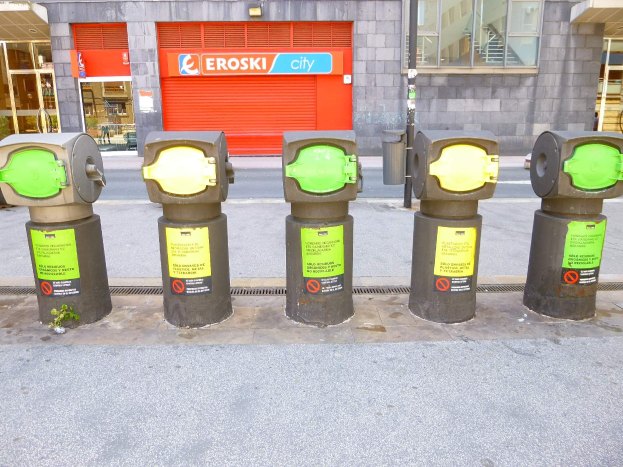 Eine Reihe von Parkautomaten entlang einer Straße mit einem Gebäude, das Glasfenster und eine Rolllade mit Text sowie einen nahen Müllcontainer zeigt.