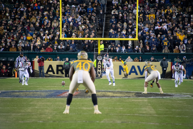 Fußballspieler in Sportkleidung und Helmen konkurrieren auf einem Feld mit Zuschauern im Hintergrund, umgeben von Stadionstrukturen einschließlich Pfosten und Tafeln mit Text.