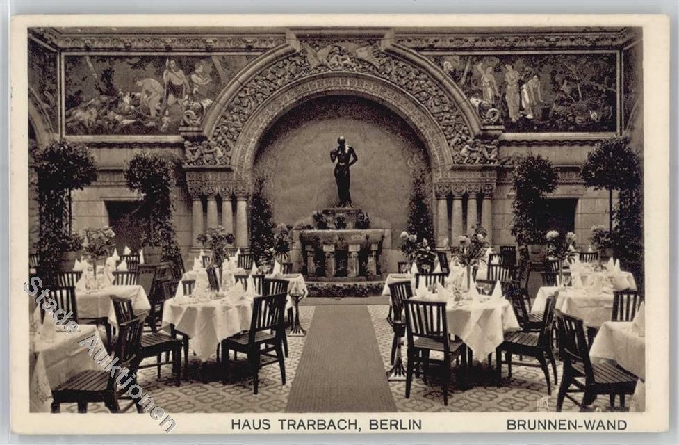 Schwarze und weiße Fotografie eines alten Berliner Restaurantinnenraums mit Tischen, Stühlen, Blumenvasen, einer Statue im Hintergrund, Wandskulpturen, Pflanzen und Text unten.