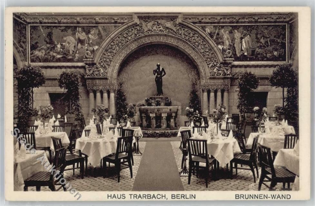 Schwarze und weiße Fotografie eines alten Berliner Restaurantinnenraums mit Tischen, Stühlen, Blumenvasen, einer Statue im Hintergrund, Wandskulpturen, Pflanzen und Text unten.