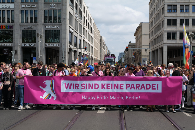Eine Gruppe von Menschen marschiert auf einer Straße in Berlin mit einem pinken Banner, auf dem "Happy Pride March" steht, mit Gebäuden, Laternenmasten und Verkehrszeichen an der Straße unter einem bewölkten Himmel.