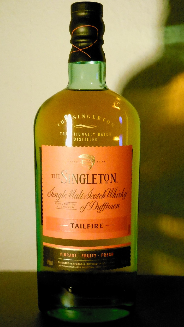 Eine tiefe bernsteinfarbene Flasche Singleton of Dufftown Tailfire Single Malt Scotch Whisky mit einem schwarzen Etikett, das eine schlagkräftige Schrift und ein Wappen mit einem Löwen und einer Burg zeigt.
