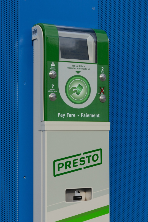 Ein grün-weißer Parkscheinautomat mit Text und Knöpfen darauf, montiert an einer blauen Wand, mit einem Wasserzeichen im rechten unteren Eck.