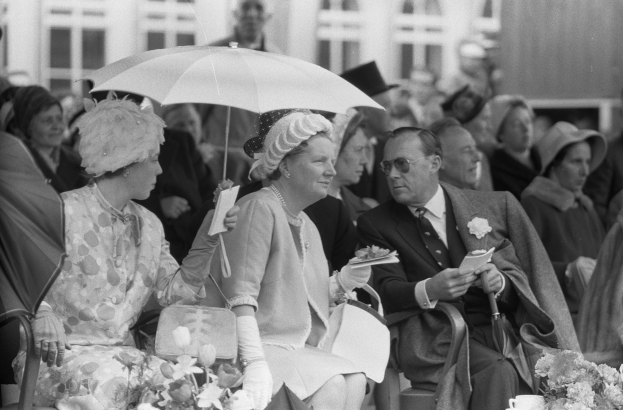 Schwarz-weiß-Foto von Queen Elizabeth II. und Prinz Philip, Duke of Edinburgh, sitzend mit Schirmen, umgeben von einer Menge mit Hüten und Büchern, mit einem Gebäude und Blumenschmuck im Hintergrund.