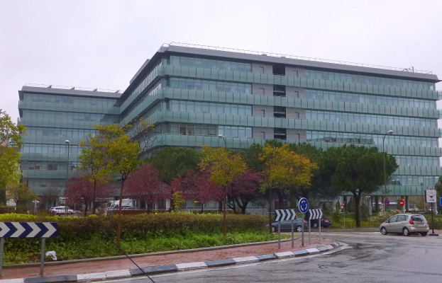 Großes modernes Gebäude mit zahlreichen Fenstern, identifiziert als Hauptquartier der Europäischen Zentralbank, umgeben von Grünflächen, Straßeninfrastruktur, fahrenden Fahrzeugen und einem bewölkten Himmel.