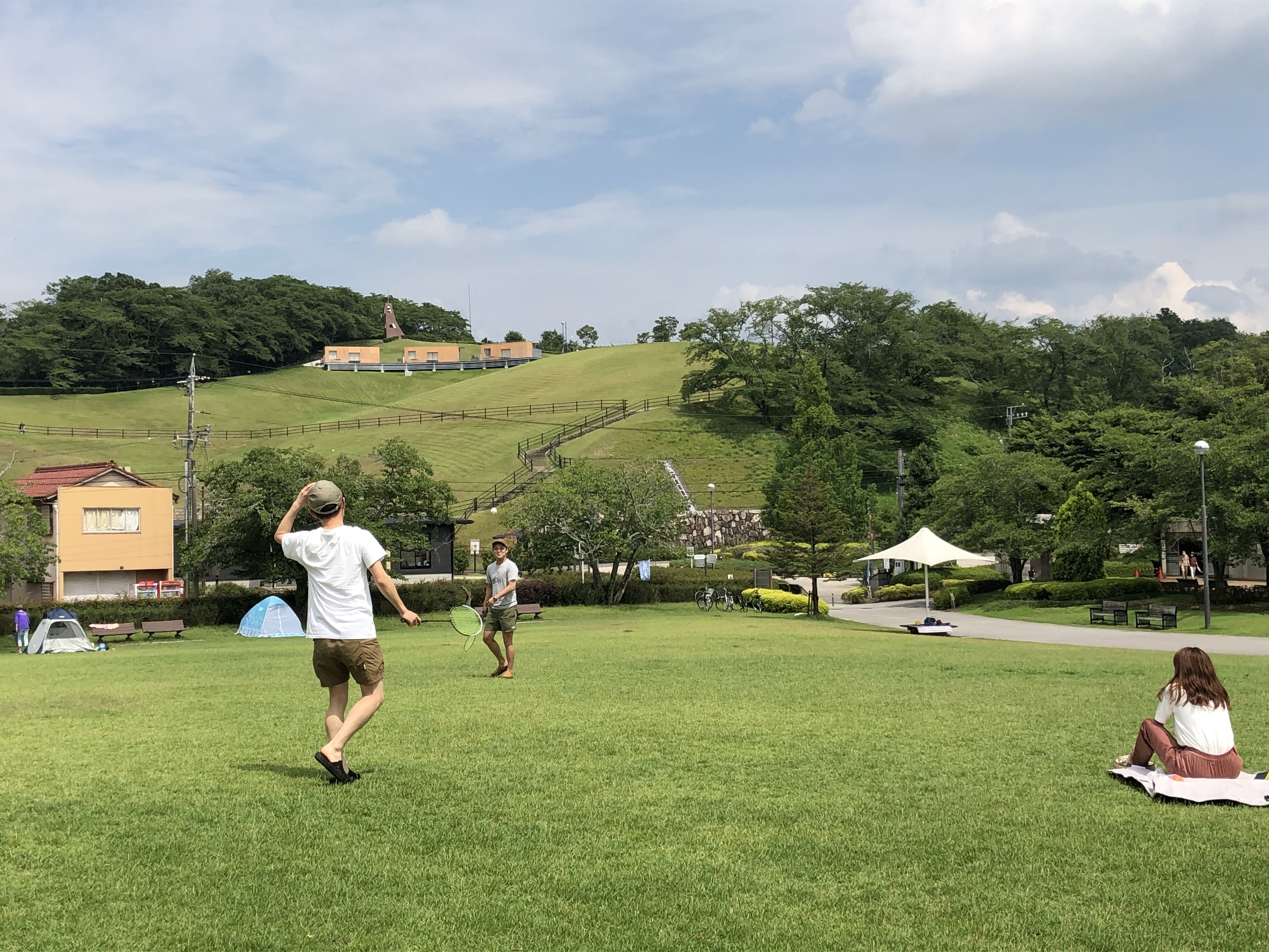 Gruppe von Menschen, die Badminton in einem Park mit Zelten, Straßenlaternen, Strommasten, Stromkabeln, Gebäuden, Bäumen, Hügeln und einem bewölkten Himmel im Hintergrund spielen.
