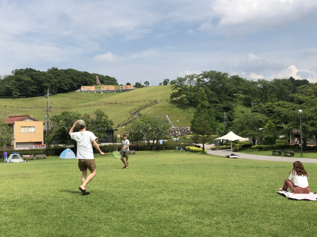 Gruppe von Menschen, die Badminton in einem Park mit Zelten, Straßenlaternen, Strommasten, Stromkabeln, Gebäuden, Bäumen, Hügeln und einem bewölkten Himmel im Hintergrund spielen.