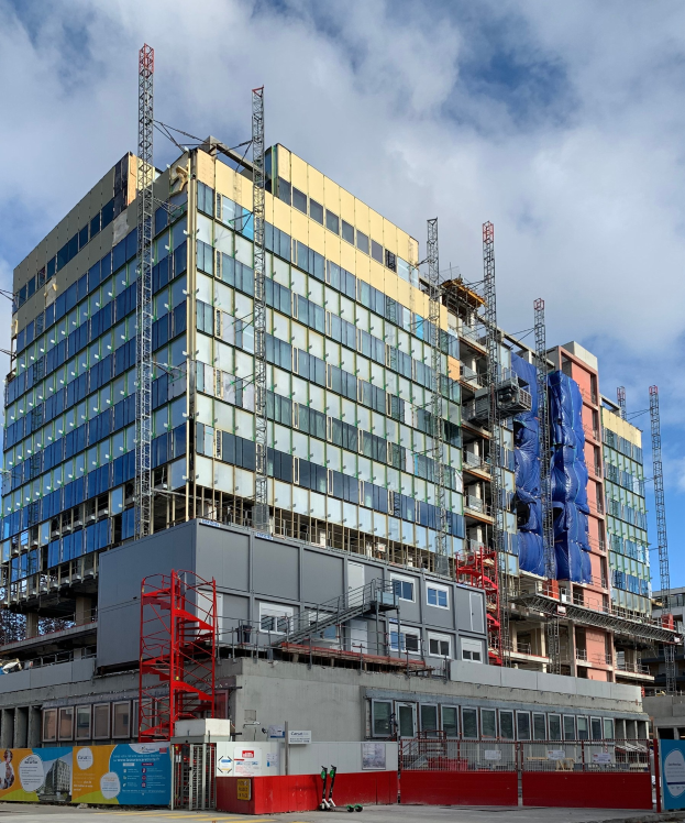 Großes Gebäude im Bau in einer städtischen Umgebung, mit Glasfenstern, Metallrahmen, einer Treppe, einem Zaun, einem Schild, Strommasten, einer Straßenlaterne, nahen Bäumen und einem bewölkten Himmel.