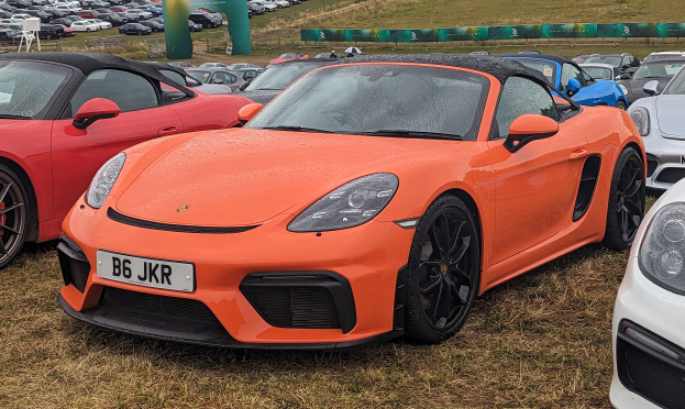 Roter Porsche 718 Boxster GTS beim Goodwood Festival of Speed, umgeben von anderen Autos auf dem Rasen, mit einem Banner, Bäumen und einem klaren blauen Himmel im Hintergrund.