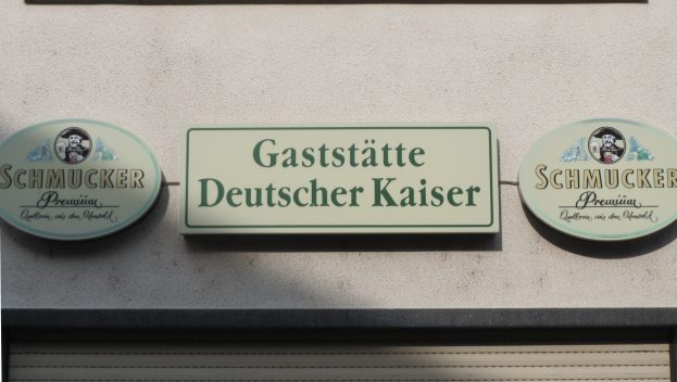 Drei hölzerne Schilder mit der Aufschrift "Gaststätte Deutscher Kaiser Schmucker" sind an einer Gebäudewand über einer geschlossenen Jalousie angebracht.