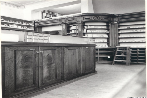 Ein Schwarz-Weiß-Foto eines Apothekeninneren mit einem Tresen, Schränken, einer Leiter und Regalen, die mit verschiedenen Gegenständen gefüllt sind, mit Text oben drauf.
