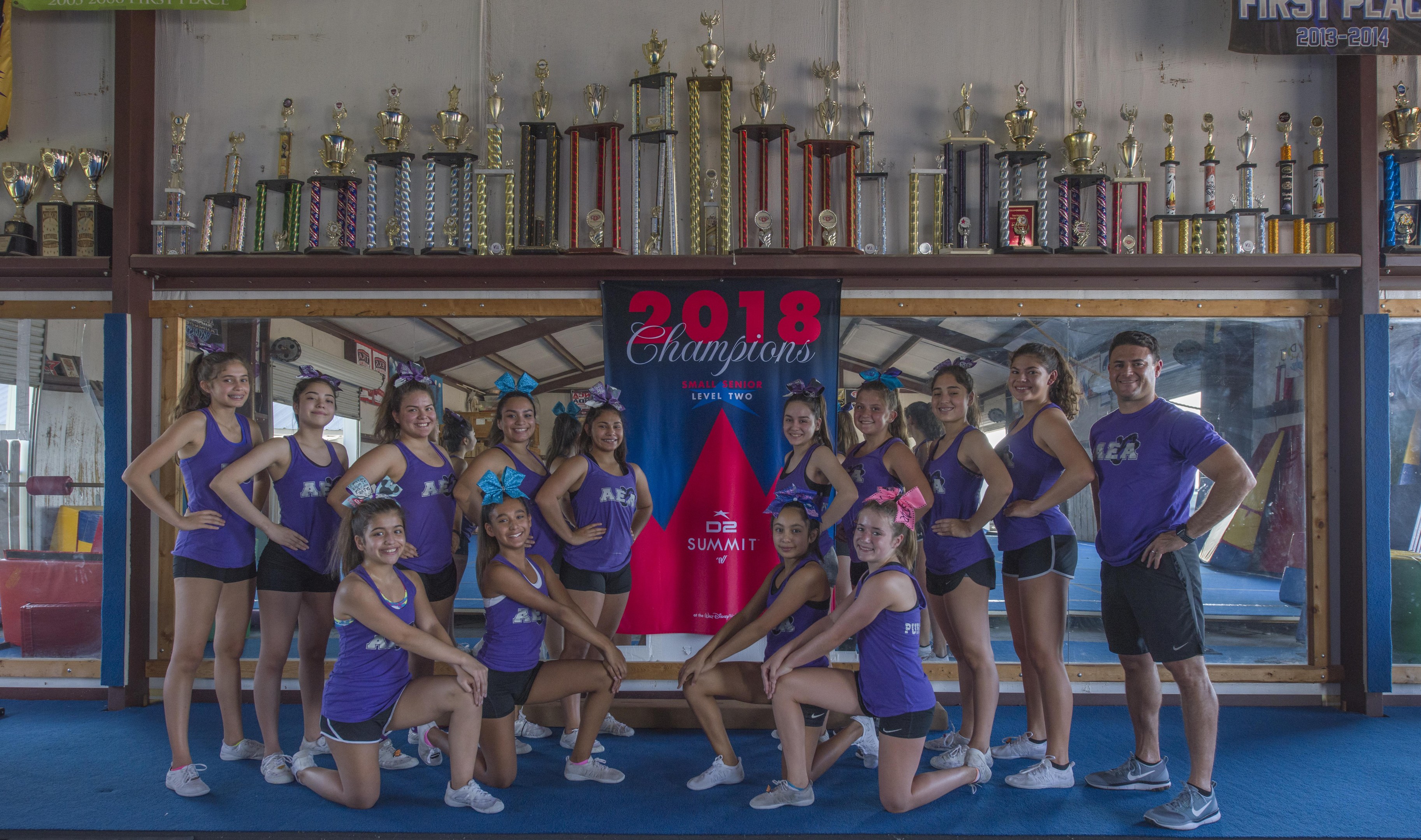 Gruppe von Cheerleadern in lila T-Shirts und schwarzen Shorts, die vor einem Trophy-Schaukasten mit einem Banner "2018 Summit Cheerleading Champions" posieren.