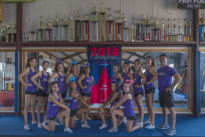 Gruppe von Cheerleadern in lila T-Shirts und schwarzen Shorts, die vor einem Trophy-Schaukasten mit einem Banner "2018 Summit Cheerleading Champions" posieren.