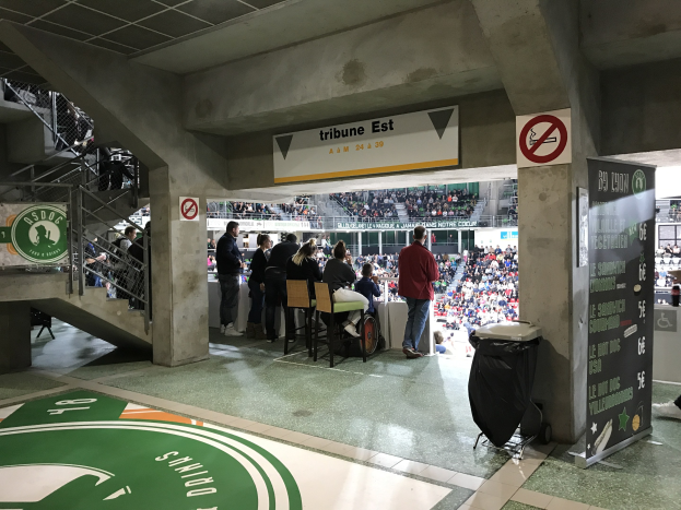 Eine Gruppe von Menschen steht vor einem sitzenden Publikum in einem Stadion, mit Schildern, die "Boston Celtics vs New York Jets" lesen, einem Müllcontainer, einer Tafel mit Text, einer Treppe mit Geländern und Deckenlampen.