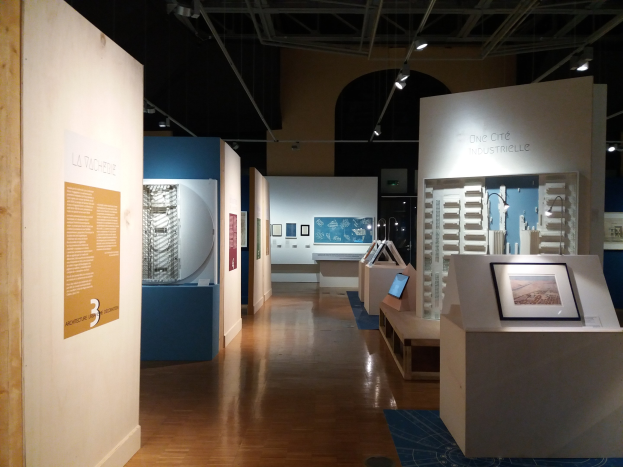 Ein Raum mit verschiedenen Kunstwerken im Museum of Modern Art, darunter gerahmte Fotos auf Tischen, Texttafeln an den Wänden, Deckenleuchten und ein Teppichboden.
